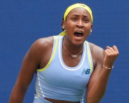 Coco Gauff celebrująca zwycięstwo lub zdobycie punktu na korcie tenisowym, z uniesionymi rękami i radosnym wyrazem twarzy, w tle rozmazana publiczność.
