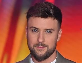 Aleksander Sikora, dziennikarz i prezenter telewizyjny, uśmiecha się, przeglądając swój telefon, w tle rozmyte studio telewizyjne lub nowoczesne wnętrze.