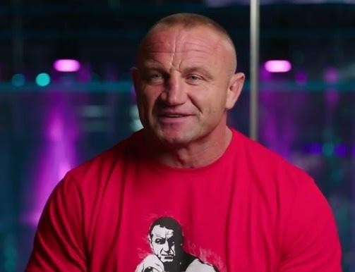 Mariusz Pudzianowski prezentujący swoją siłę, w akcji strongman lub w dynamicznej pozie MMA, z widoczną muskulaturą i skupionym wyrazem twarzy.